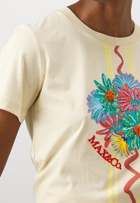 Camiseta de algodón crema con bordado floral colorido, texto rojo "MAX&Co." y dos franjas amarillas en ondulación a los lados. Cuello redondo, mangas cortas.