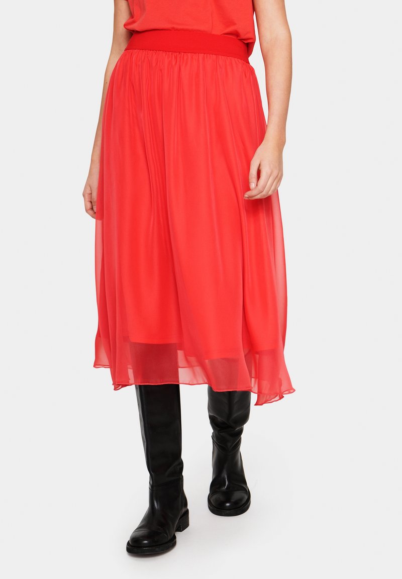 Personne portant une jupe midi fluide rouge vif à large ceinture, un haut rouge assorti et des bottes noires montant jusqu’aux genoux, debout.