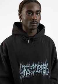 Black hoodie featuring "WASTED" in blue flame-like lettering. Textured material with a drawstring hood and silver tips on the drawstrings.