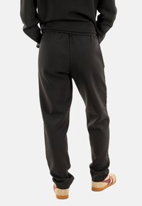Pantalones de chándal negros con cintura elástica y tobillos ajustados, hechos de una tela suave; llevados con zuecos beige con acentos color burdeos.