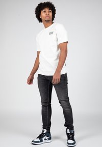 Bílý tričko s krátkým rukávem a černým logem na levé hrudi, spárované s černými slim-fit džíny a modro-bílými teniskami.