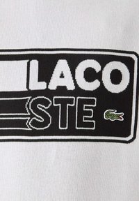 Czarnobiały prostokątny naszywka z logo marki z napisem "LACOSTE" i małym zielonym krokodylem na białym tle.