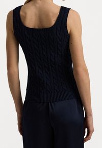Kvinna som bär en ärmlös marinblå kabelstickad tröja och mörka byxor, sedd bakifrån mot en enkel ljus bakgrund.