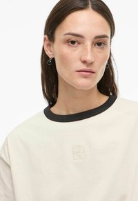 Mlada ženska z bledimi pegami, ki nosi črne obroče za ušesa in kremno majico s črnim ovratnikom ter majhnim vezenim logotipom.