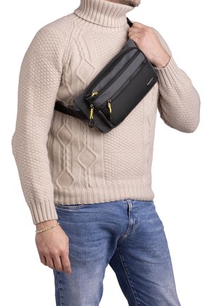 Sac bandoulière noir avec deux compartiments zippés, tirettes jaunes et surface texturée, porté au-dessus d'un pull en maille beige.