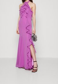 Long purple dress with ruffles along the neckline and side seam. Features a high slit and is paired with black strappy heels and a small bag.