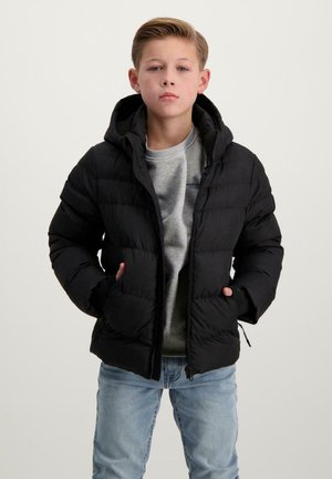 Een jongen staat zelfverzekerd, gekleed in een zwarte gewatteerde jas over een grijze hoodie en blauwe jeans, met zijn handen in zijn zakken.