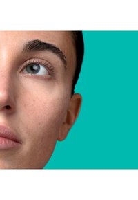 Zbliżenie twarzy o jasnej karnacji, wyraźnych brwiach i czystym niebiesko-szarym oku, na jednolitym turkusowym tle.