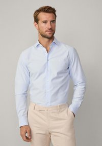 Lichtblauwe overhemd met lange mouwen en een button-down kraag, gladde textuur en een klein logo op de borst, gecombineerd met beige broek.