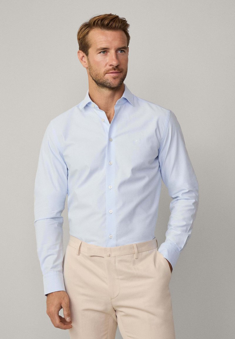 Lichtblauwe overhemd met lange mouwen en een button-down kraag, gladde textuur en een klein logo op de borst, gecombineerd met beige broek.