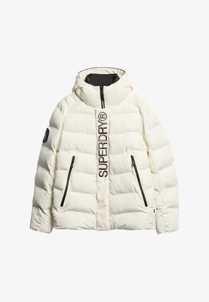 Chaqueta acolchada en crema con cuello alto, cierre de cremallera y dos bolsillos laterales. Presenta texto de logo negro y acentos en el borde. Textura suave y acolchada.