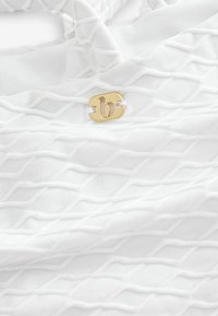 Tessuto bianco con un motivo a diamante testurizzato, con un'etichetta in metallo dorato attaccata da un filo bianco. Materiale liscio e leggero.
