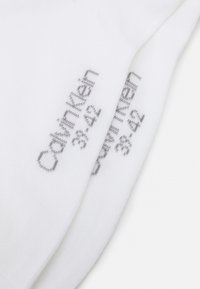 Calzini bianchi in cotone con testo grigio che riporta "Calvin Klein" e taglie scarpa 38-42. Texture liscia e design standard di lunghezza normale.