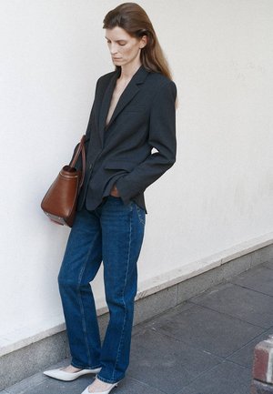 Femme en blazer sombre, jean bleu et chaussures blanches pointues, debout contre un mur blanc tenant un sac à main marron.
