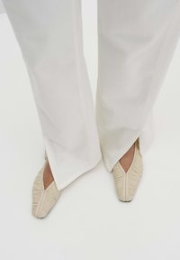 Beige ballet flats with gathered texture, pointed toe, and slight heel, worn with white wide-leg pants featuring side slits.