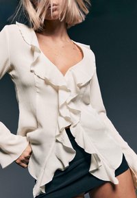 Weißes Blusenshirt mit fransigen Rändern, strukturiertem Stoff, V-Ausschnitt und Knopfverschlüssen. Kombiniert mit einem schwarzen Rock mit ausgestellt geschnittenen Ärmeln.