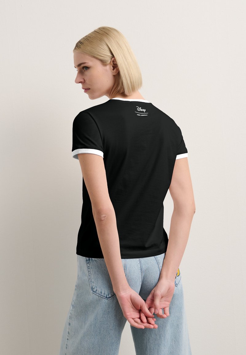 Czarna koszulka z białym kołnierzykiem i mankietami, z małym logo Disney na plecach, wykonana z miękkiego materiału, zestawiona z jasnoniebieskimi dżinsami.