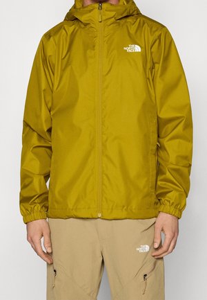 Homme portant une veste à capuche jaune moutarde et un pantalon beige, tous deux avec des logos blancs The North Face, debout avec les bras détendus.