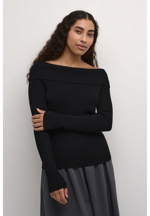 Zwarte off-the-shoulder geribde trui met lange mouwen en duimopeningen, gecombineerd met een vloeiende grijze rok, tegen een neutrale achtergrond.