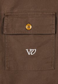 Smeđi tkaninski džep s kopčom od zlatnog dugmeta i bijelim izvezenim logotipom "VØ." Teksturirani platneni materijal.