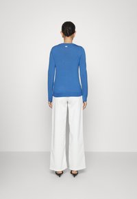 Mujer con cabello oscuro recogido en un moño, vistiendo una blusa azul de manga larga, pantalones blancos de pierna ancha y sandalias negras de tacón, de espaldas en un fondo liso.