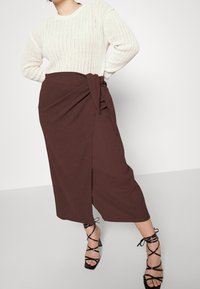 Crème gebreide trui en een bruine wraprok tot de knie, met een zijstrik. Zwarte sandalen met hakken en bandjes maken de outfit compleet.