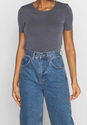 Femme portant un t-shirt gris à manches courtes ajusté, rentré dans un jean en denim bleu taille haute, debout devant un fond uni.