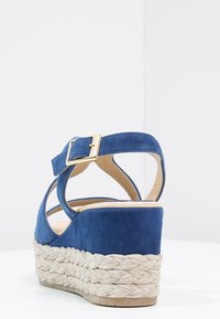 Sandalo con zeppa in suede blu, suola in corda di juta e cinturino regolabile dorato. I fori laterali arricchiscono il design, offrendo un tocco moderno.