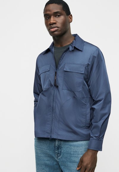 Homme portant une veste zippée bleu marine avec des poches sur la poitrine, par-dessus une chemise noire et un jean bleu, debout devant un fond blanc uni.