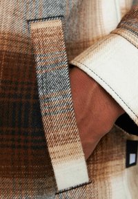 Chaqueta de lana con un patrón de cuadros en marrón, crema y azul. Presenta un cuello blanco y un bolsillo lateral con detalles de costura visibles.