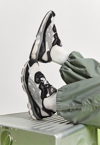 Schwarz-silberne Sportschuhe mit Netzpaneelen, strukturierten Gummisohlen und schwarzen Schnürsenkeln, getragen mit weißen Socken und lockeren grünen Hosen.