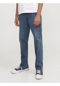 Persona vestida con jeans rectos azules, camiseta blanca y zapatillas azul marino con suelas y cordones blancos, de pie sobre un fondo liso.