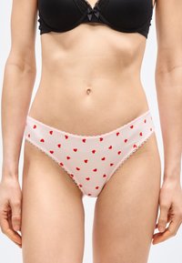 Γυναίκα που φοράει μαύρο σουτιέν και ανοιχτό ροζ εσώρουχο με κόκκινα σχέδια καρδιάς, φωτογραφημένη από τη μέση μέχρι το μέσο του μηρού σε ουδέτερο φόντο.