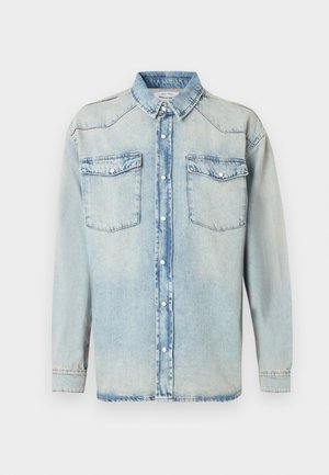 Camisa de mezclilla azul claro con mangas largas, dos bolsillos en el pecho, cierre de botones y cuello clásico. Textura desgastada por todo el tejido.