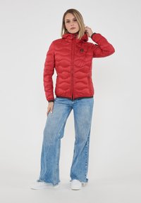 Donna con giacca trapuntata rossa con cappuccio, jeans a gamba larga blu chiaro e sneakers bianche, in piedi su uno sfondo bianco semplice.