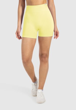 Frau trägt gelbe Sportshorts mit hoher Taille und weiße Sneakers, steht mit einem Bein leicht nach vorne vor einem schlichten Hintergrund.