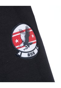 Zwarte stof heeft een ronde patch met een adelaarontwerp in grijs en rood, omrand door een zwarte cirkel, vergezeld van twee rode strepen en de tekst "BOB."