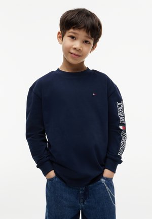 Dreng iført en marineblå langærmet skjorte med teksten "TEAM" på ærmet og blå jeans, stående med hænderne i lommerne mod en hvid baggrund.