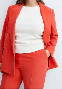 Blazer ajusté orange vif sur un haut blanc à côtes. Le blazer présente des épaules structurées et un seul bouton, avec un pantalon assorti.