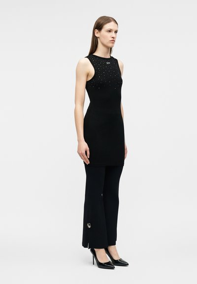 Γυναίκα με μαύρο μίνι φόρεμα χωρίς μανίκια με μικρές διακοσμήσεις, συνδυασμένο με μαύρο παντελόνι με καμπάνα και μαύρα ψηλοτάκουνα παπούτσια, στέκεται πλευρικά.