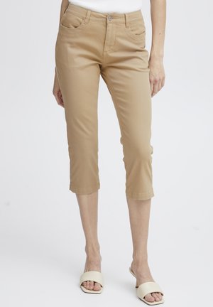 Pantalons capri beiges en mélange de coton doux, avec une coupe slim, des poches avant et des ourlets repliés. Associés à des sandales de couleur claire.