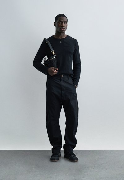 Άνδρας ντυμένος με μαύρη μακρυμάνικη μπλούζα, φαρδιά μαύρα παντελόνια, μαύρα παπούτσια, κρατώντας μικρή μαύρη τσάντα ώμου, στέκεται μπροστά σε απλό ανοιχτό γκρι φόντο.