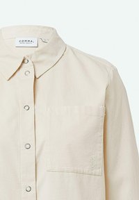 Camicia beige chiaro con chiusura a bottoni, colletto a punta e tasca sul petto; presenta bottoni a contrasto e un tessuto texturizzato.