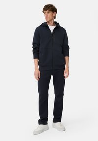 Marineblå zip-up hoodie med justerbar snor i hætten, frontlommer og matchende marineblå bukser. Bæres med hvide sneakers. Glat tekstur.