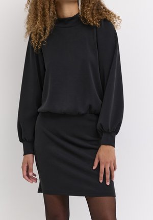 Kobieta ubrana w czarną sukienkę z długim rękawem, bufkami na ramionach i dopasowaną spódnicą, stojąca na jasnym, jednolitym tle.