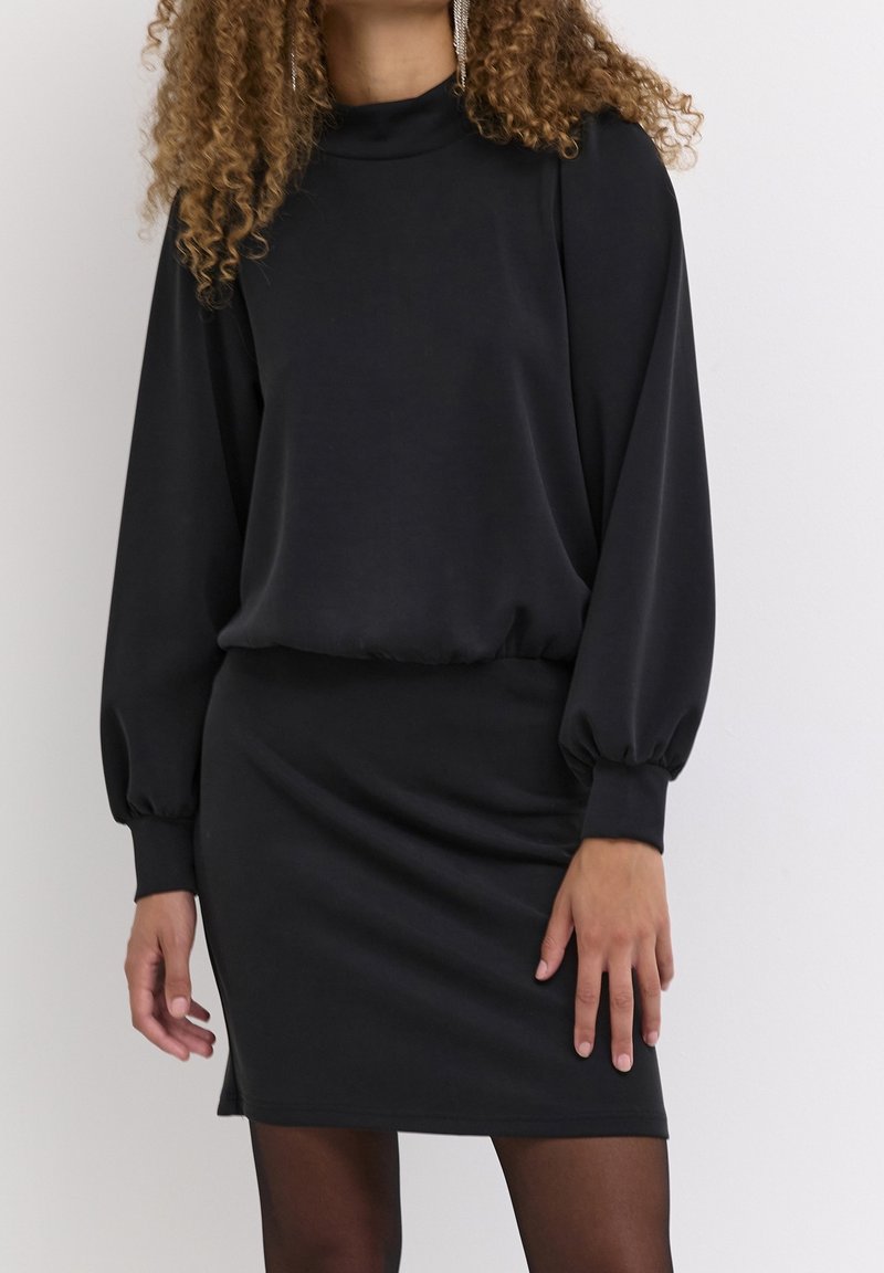 Femme portant une robe noire à manches longues bouffantes et jupe ajustée, debout devant un fond clair uni.