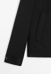 Schwarzer Wollmischmantel mit strukturiertem Design, umgeschlagenem Ärmel mit Knopfdetail und einer diskreten Seitentasche.