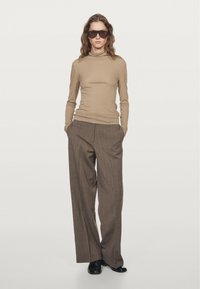 Beige turtleneck top paired with brown pinstripe wide-leg trousers. Model stands with hands in pockets, wearing black sunglasses and shoes.