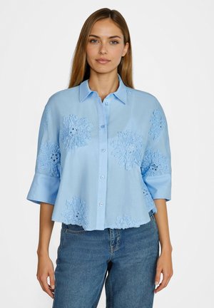 Junge Frau trägt eine hellblaue Bluse mit Knöpfen, bestickt mit floralen Mustern und 3/4-Ärmeln, kombiniert mit blauen Jeans.