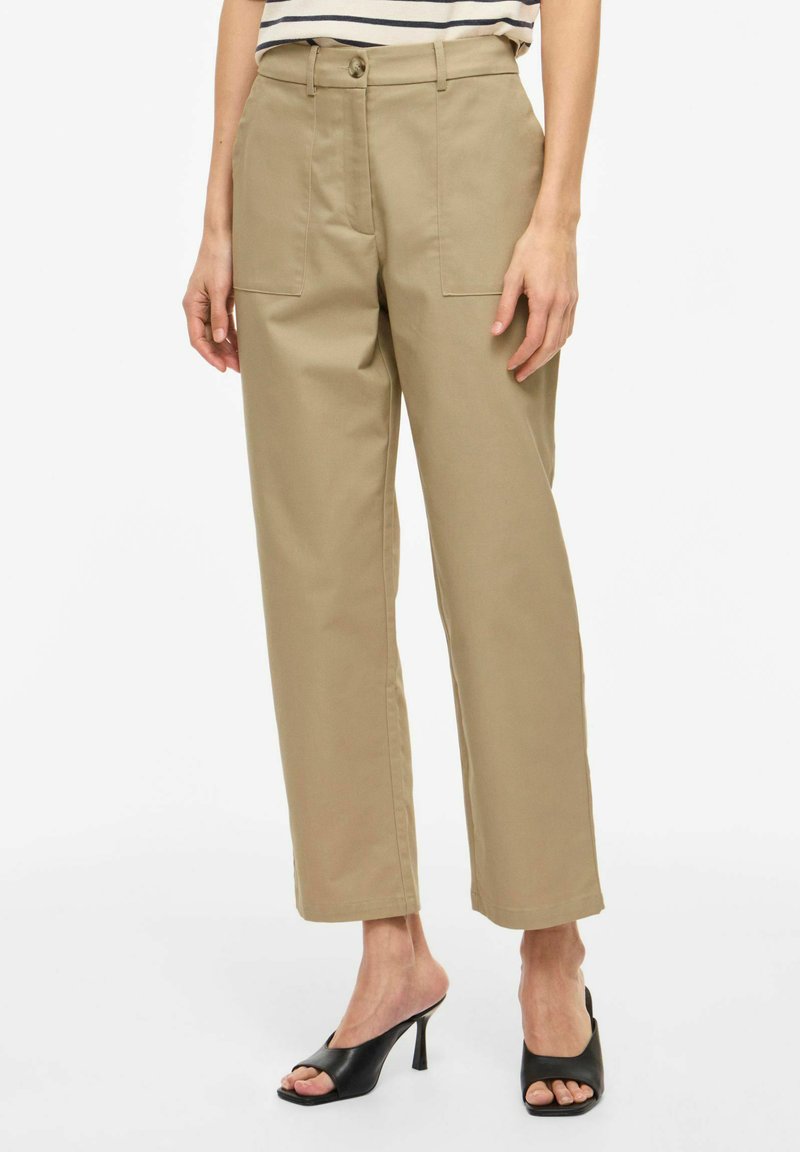 Femme portant un pantalon beige à jambes droites avec des poches plaquées à l'avant et des mules à talons ouvertes noires, debout devant un fond blanc.